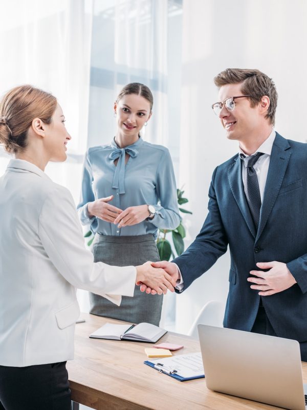 cheerful recruiter shaking hands with woman near colleague in office